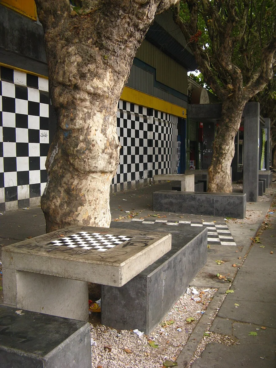 Mesas de hormigón con tableros de ajedrez bajo los árboles, frente a un muro pintado a cuadros blancos y negros.
